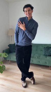 Blauwe trui met rits, zwarte broek en bruine schoenen. Staat in een kamer met een groene bank en een lamp, met een plant op de voorgrond.