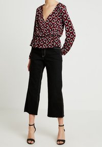 Top avvolgente a fantasia rossa e nera con scollatura a V profonda e vita elastica, abbinato a pantaloni neri a tre quarti e cinturini alla caviglia.