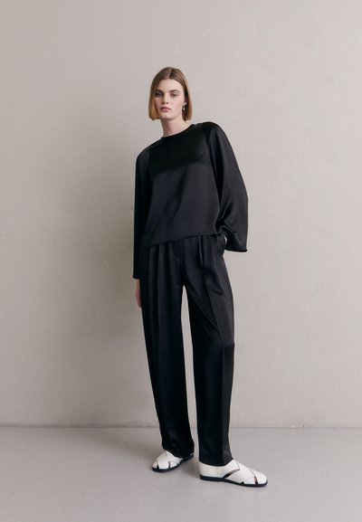 Haut en satin noir ample avec des manches larges, associé à un pantalon large assorti. Le mannequin porte des sandales blanches avec des accents noirs.