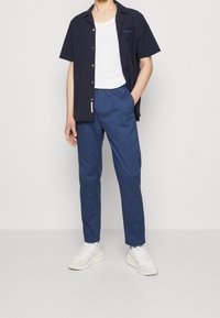 Marineblauwe short-sleeve shirt met knopen, witte tanktop eronder, blauwe taps toelopende broek en witte sneakers. Schone lijnen, gladde stoffen.