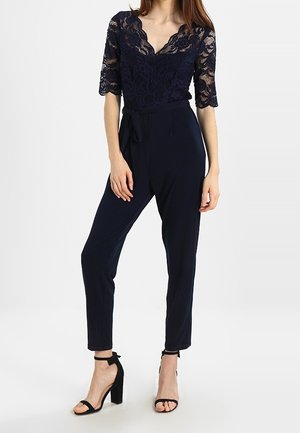 Donna che indossa un top blu navy in pizzo con scollo a V e maniche a 3/4, pantaloni blu navy a vita alta con cintura annodata e sandali con tacco a blocco e cinturino alla caviglia neri.