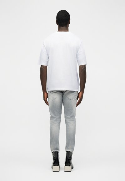 Άνδρας που κοιτάει προς τα πίσω, φοράει ένα απλό λευκό κοντομάνικο μπλουζάκι, ανοιχτό γκρι τζιν και μαύρα μποτάκια αστραγάλου με ψηλό τακούνι.