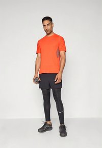 Helekollane spordisärk halli aktsendiga, koos mustade lühikeste pükste ja mustade retuusidega. Modell hoiab ühes käes hantlit.