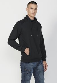 Sudadera negra con cuello alto y cordones, con un patrón de espiga, bolsillos laterales y puños acanalados. Combinada con pantalones cargo azules.