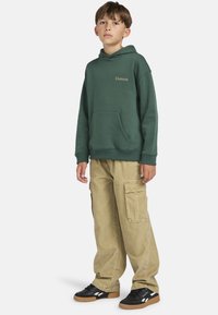 Junge trägt einen grünen Element-Hoodie, beigefarbene Cargo-Hosen und schwarze Reebok-Sneaker und steht vor einem schlichten hellen Hintergrund.