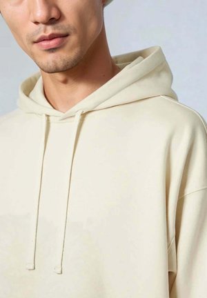 Sudadera con capucha color crema y corte relajado, con cordones y tela suave y texturizada. Diseño simple, sin patrones visibles.