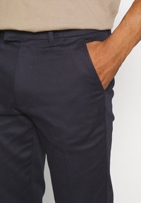 Granatowe spodnie w kant o gładkiej fakturze, z przednimi kieszeniami i dopasowanym krojem, zestawione z jasnobrązową bluzką widoczną powyżej.