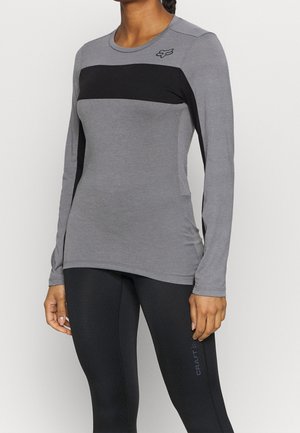 Vrouw die een nauwsluitend grijs shirt met lange mouwen draagt met een zwarte horizontale streep en een klein logo op de bovenborst, gecombineerd met zwarte legging.