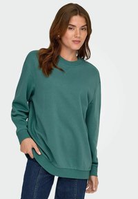Felpa color teal in un morbido tessuto con scollo rotondo, polsini e orlo a coste. Abbinata a jeans di denim per un look rilassato.