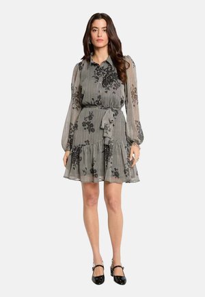 Femme portant une robe longue à manches longues à motifs avec des dessins floraux noirs, cintrée à la taille, et des chaussures Mary Jane noires, debout devant un fond uni.