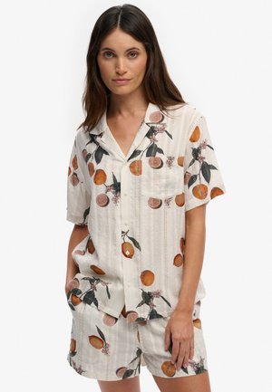 Femme portant une chemise blanche à manches courtes boutonnée et un short assorti avec un imprimé de fruits orange et feuilles vertes, debout avec une main dans la poche.