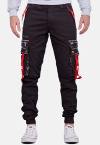 Schwarze Cargo-Hosen mit roten Akzenten, mehreren Taschen, Reißverschlussdetails und Nietenverzierungen. Umschläge an den Knöcheln und strapazierfähiger Stoff.