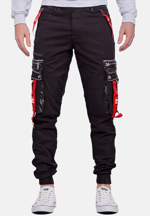 Pantaloni cargo neri con dettagli rossi, numerose tasche, particolari con cerniera e decorazioni con borchie. Caviglie risvoltate e tessuto resistente.