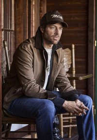 Homme en veste marron, jeans bleus et casquette noire assis sur une chaise en bois à l'intérieur, regardant pensivement sur le côté avec les mains jointes.