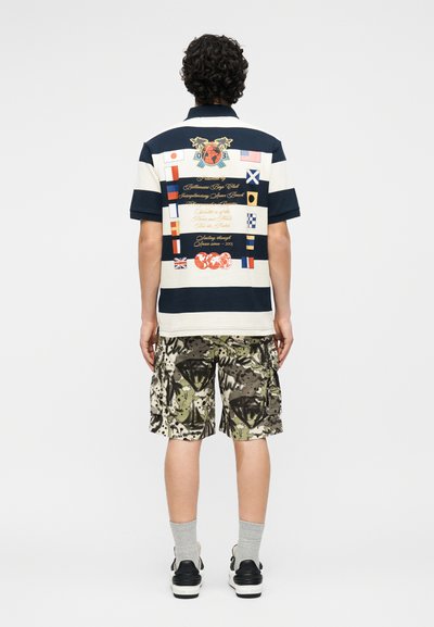 Mann trägt ein marineblau-weiß gestreiftes Poloshirt mit nautischen Flaggen und Schriftzug auf dem Rücken, Camouflage-Cargoshorts, graue Socken und schwarze Turnschuhe.