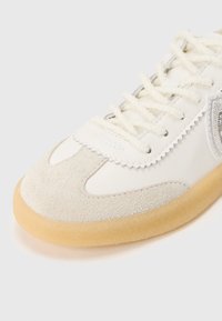 Baltas odinis batas, turintis pilką zomšos pirštų dangtelį, tekstūruotus sidabro akcentus ir kreminės spalvos raištelius, viskas ant smėlio spalvos gumos pado.