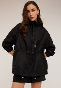 Svart lättviktig jacka med huva, elastiska ärmslut, dragsko i midjan och metalliska detaljer. Tillverkad av slätt, glänsande tyg.