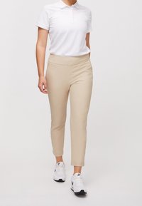 Béžové slim-fit kalhoty s hladkým povrchem, se širokým pasem a bočními kapsami, v kombinaci s jednobarevným bílým polo tričkem a bílými teniskami.