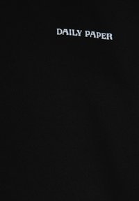 Zwarte katoen stof met witte afgedrukte tekst die "DAILY PAPER" zegt in een gestileerd lettertype nabij de bovenste linkerhoek.