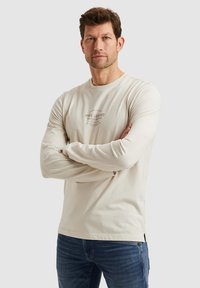 Lys beige langærmet t-shirt i blød bomuld med rund hals og et grafisk logo på forsiden. Sammen med mørke denimjeans.