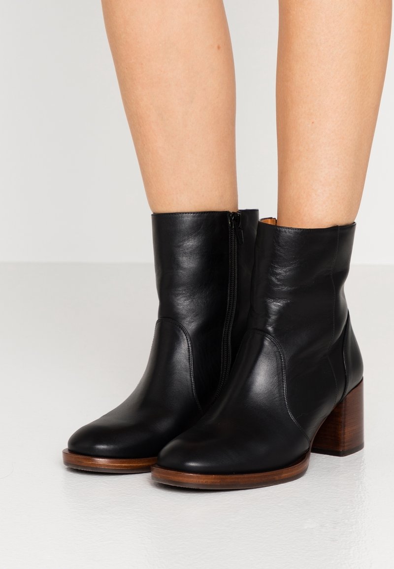 Botines de cuero negro con cremallera lateral, punta redonda y tacón cuadrado de madera marrón. Textura lisa y diseño ajustado.