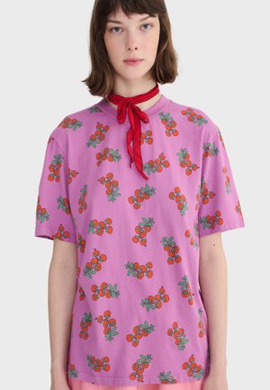 Giovane che indossa una maglietta viola con stampe di ciliegie rosse e un fazzoletto rosso annodato a fiocco su uno sfondo semplice.