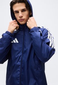 Νεαρός άνδρας που τραβάει τη κουκούλα ενός μπλε αθλητικού μπουφάν Adidas με λευκές ρίγες στα μανίκια, σε απλό φόντο.