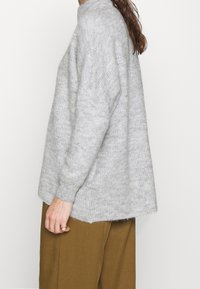 Šedý pletený svetr s vysokým límcem, žebrovanou strukturou, sklopenými rameny a uvolněným střihem. V kombinaci s hnědými kalhotami s širokými nohavicemi.