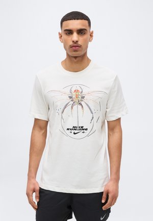 Tânăr purtând un tricou alb Nike Running cu un grafic mare stilizat cu o insectă, asortat cu pantaloni scurți negri pe un fundal simplu.