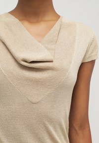 Zbliżenie osoby noszącej beżowy sweter z krótkim rękawem i drapowanym dekoltem typu komin.