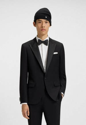 Svart tuxedo med satinrevers, vit skjorta, svart fluga och en svart stickad mössa. Bröstficka ger kontrast. Skräddarsydd passform.