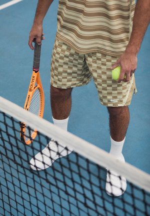 Tennisspeler in geruite korte broek en gestreept shirt staat achter het net met een oranje racket in de linkerhand en een gele bal in de rechterhand.