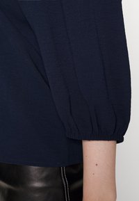 Top blu navy con una texture morbida, caratterizzato da maniche a sbuffo con polsini elasticizzati. L'orlo è leggermente curvato, completando il design.