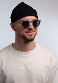 Occhiali da sole grigi con lenti rotonde, accompagnati da un berretto lavorato a maglia nero. Il soggetto indossa una maglietta chiara a collo rotondo.