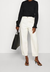 Blusa nera a maniche lunghe con scollatura rotonda e polsini a volant, abbinata a pantaloni bianchi a gamba larga e borsa nera. Sandali neri con tacco.