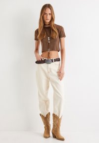 Brun cropped top med kant, beige højtaljede bukser og brune cowboystøvler med syninger. Accessories omfatter et dekorativt bælte.