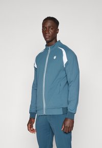 Jachetă de trening pentru bărbați cu fermoar, din material teal, cu accente albe în dungi, guler înalt și manșete ribbate. Are un logo pe piept.