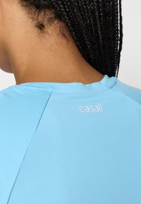 Ανοικτό μπλε αθλητικό μπλουζάκι με στρογγυλή λαιμόκοψη και λεία υφή. Διαθέτει το λογότυπο "casall" σε λευκό χρώμα στην επάνω πλάτη κοντά στη λαιμόκοψη.