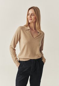 Beige V-Ausschnitt-Pullover aus weichem Stoff mit Kragen. Ausgestattet mit langen Ärmeln und einem taillierten Saum, kombiniert mit dunkelblauen Jeans.