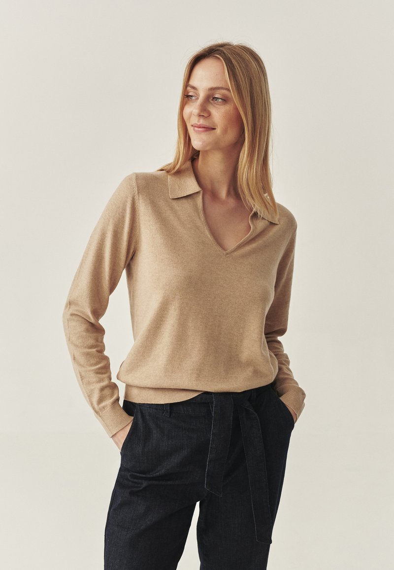 Beige V-Ausschnitt-Pullover aus weichem Stoff mit Kragen. Ausgestattet mit langen Ärmeln und einem taillierten Saum, kombiniert mit dunkelblauen Jeans.