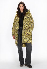 Puffjacka med gul bas, svart och lila leopardmönster, med huva och stora framfickor, kombinerad med svarta jeans och stövlar.