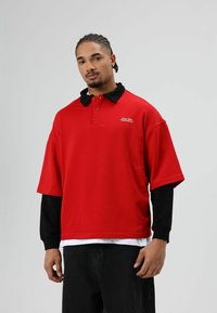 Rood polohemd met korte mouwen en een zwarte kraag, met een logodetail. Draagt over een zwart shirt met lange mouwen en zwarte broek.