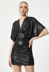 Zwarte glanzende jurk met een diepe V-hals, korte mouwen en geraffineerde details bij de taille. De gladde textuur accentueert de silhoutte.