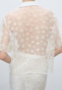 Camicia bianca trasparente con maniche corte, caratterizzata da ricami floreali in giallo e bianco. Design con colletto e una texture leggera e traslucida.