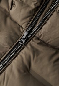 Pufferjakke sektion i dæmpet brun med en sort lynlås. Stoffet er glat med et subtilt gittermønster, der fremhæver den quiltede tekstur.