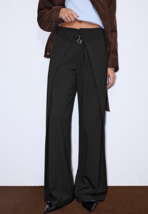 Femme portant un pantalon noir à rayures fines, taille haute, jambes larges, avec un détail de ceinture croisée et une double boucle à la taille, un haut bleu clair et une veste marron.