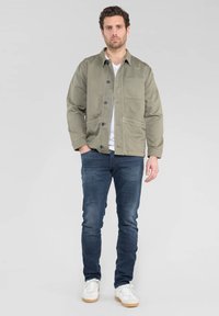 Veste en toile vert olive avec un devant à boutons, deux poches avant, portée sur un t-shirt blanc, associée à un jean bleu foncé et des baskets blanches.