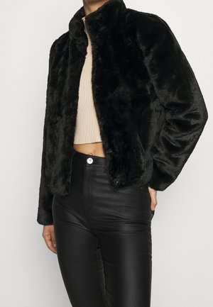 Mujer con chaqueta de piel sintética negra sobre un suéter corto de canalé beige y pantalones de cuero negro de talle alto.