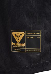 Svart tygmärke med guldtext och logotyp. Texten inkluderar "Hummel Sportswear", "Serving the Game" och "Sedan 1923", med varumärkesdetaljer.