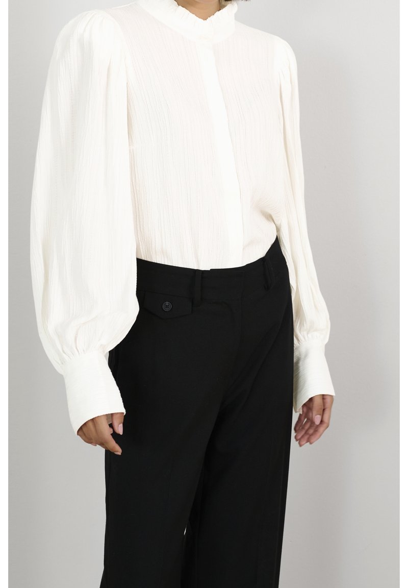 Witte, textuurgegeven blouse met een gerimpelde kraag en voluminieuze mouwen, gecombineerd met op maat gemaakte zwarte broek met zijzakken en knoopdetails.